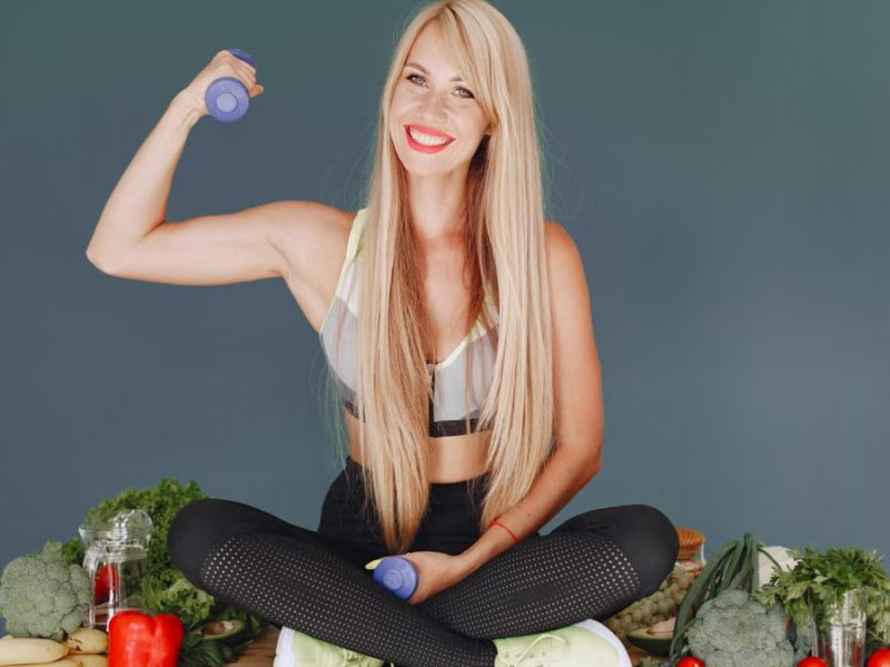 Woman lifting a small dumbbell while surrounded by fresh vegetables, embodying health and fitness.